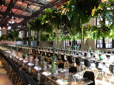 Lange gedeckte Tafeln in einer großen Halle, die Decke ist dekoriert mit tropischen Pflanzen